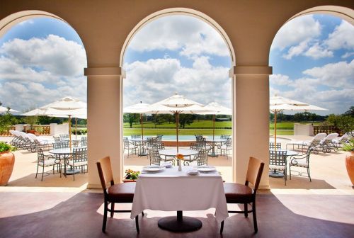 A serene outdoor dining area with a table set for two, framed by arches and overlooking a pool and lush landscape.