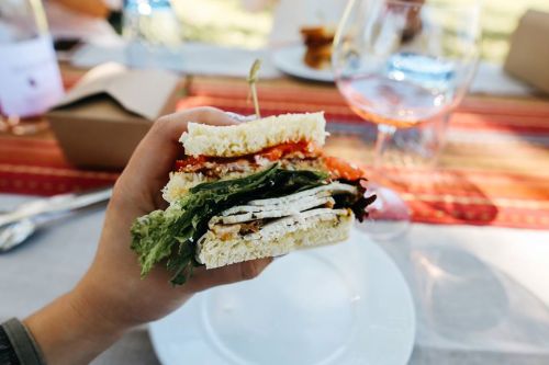 A hand holding a layered sandwich with lettuce, chicken, and tomato, with a glass of wine in the background.