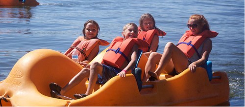 Four children wearing life jackets enjoy a sunny day on a yellow paddle boat in calm water.