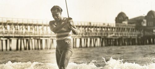 A vintage photo of a man in swimwear wading through waves near a pier, capturing a moment of seaside leisure.