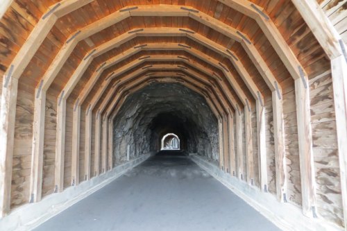 A wooden tunnel with a curved ceiling, leading into a rocky passageway, illuminated by natural light.