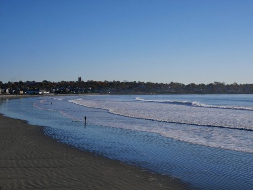 A serene beach scene with gentle waves, a person walking along the shore, and a clear blue sky in the background.