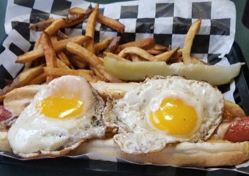 A hot dog topped with two fried eggs, served with a side of French fries and a pickle on a checkered paper-lined tray.