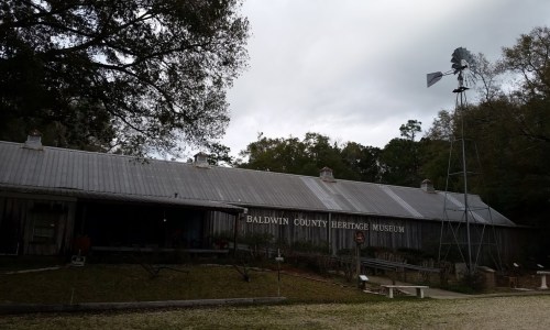 Baldwin County Heritage Museum building with a windmill nearby, surrounded by trees and grassy area.