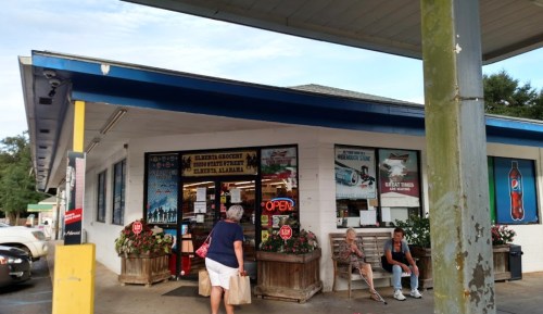 A small grocery store with an "Open" sign, featuring flower planters and two people sitting outside.