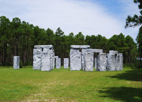 A replica of Stonehenge made of large, gray stones set in a grassy field surrounded by trees.