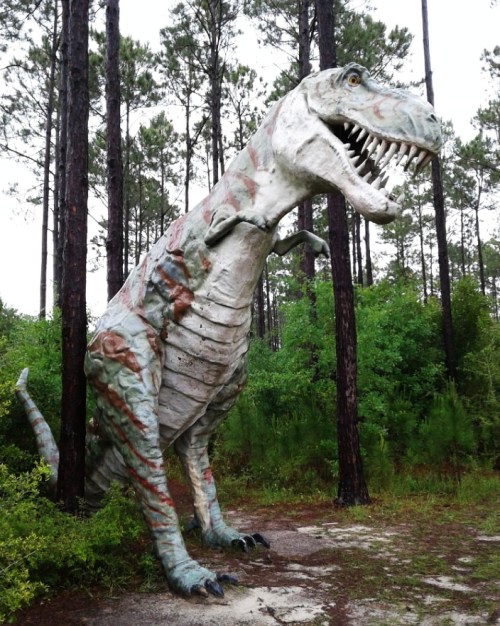 A large, painted dinosaur statue stands among tall trees and greenery in a forested area.
