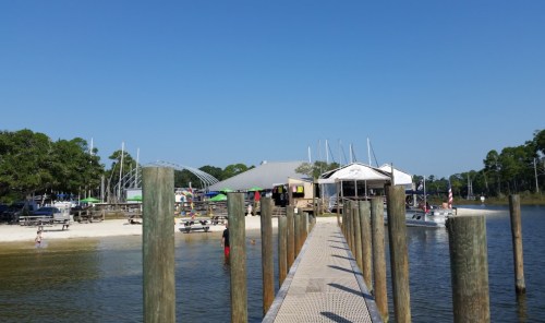A wooden dock leads to a beachside restaurant with boats and picnic areas under a clear blue sky.
