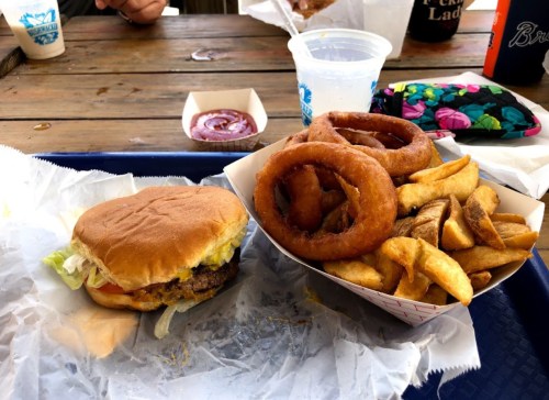 A burger with lettuce and tomato next to a basket of onion rings and fries on a tray.