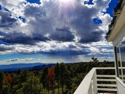 A scenic view of a cloudy sky with sunlight breaking through, overlooking a forested landscape and distant mountains.