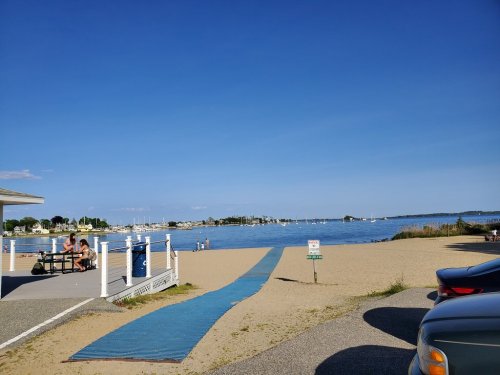 A sandy beach with a blue walkway leading to the water, boats in the distance, and a clear blue sky.