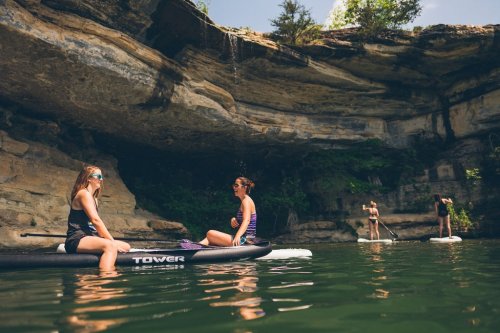 Two women sit on paddleboards in a serene, rocky cove, while two others paddle in the background under a sunny sky.