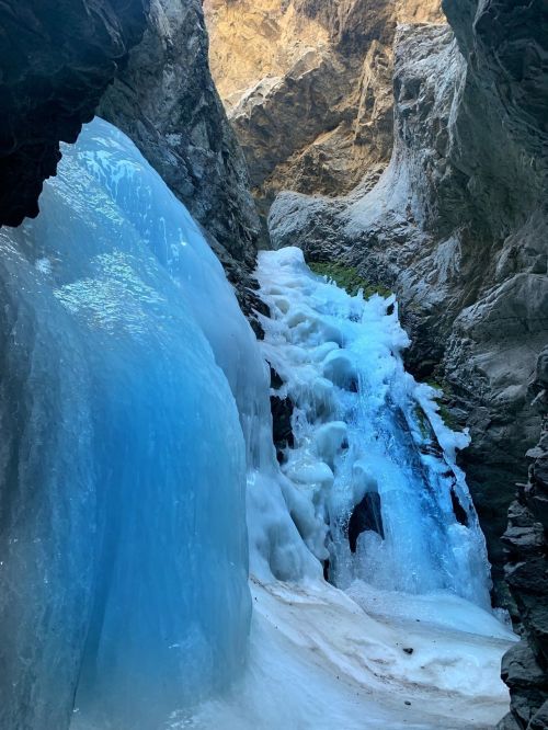 A frozen waterfall cascading down rocky cliffs, surrounded by rugged terrain and illuminated by natural light.