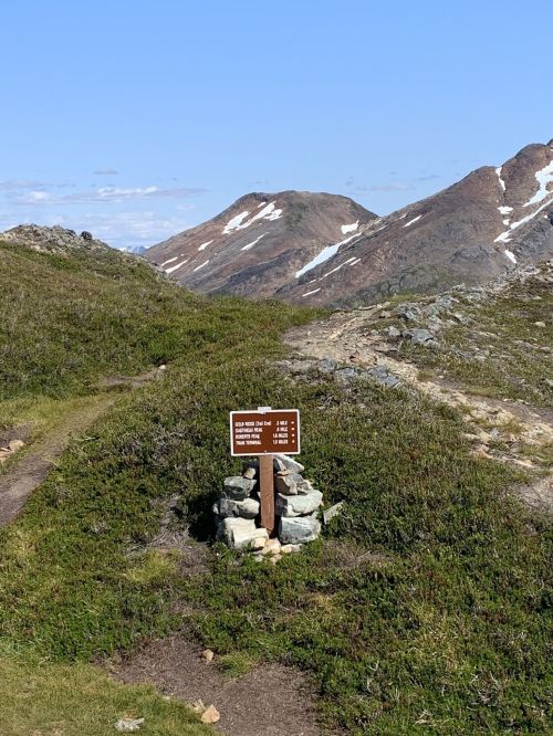 A trail sign marks a path through green terrain with rocky mountains and snow-capped peaks in the background.