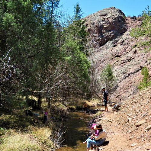 A scenic creek surrounded by trees and rocky hills, with people relaxing and enjoying nature along the water's edge.