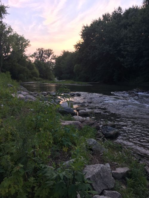 A serene riverbank at sunset, with rocks and lush greenery lining the water under a colorful sky.