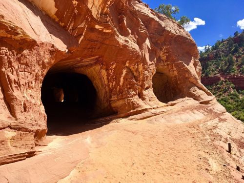Two large cave openings in a red rock formation, surrounded by a sandy path and blue sky with scattered clouds.