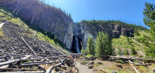 A scenic view of a waterfall surrounded by rocky terrain and tall trees under a clear blue sky. Hikers are visible in the foreground.