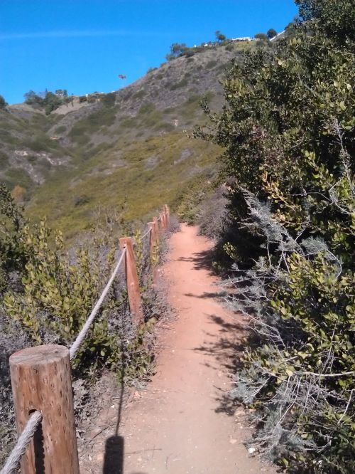 A narrow dirt path lined with wooden posts and rope, surrounded by greenery and hills under a clear blue sky.