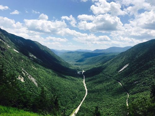 A lush green valley stretches between mountains under a partly cloudy sky, with a winding river visible below.