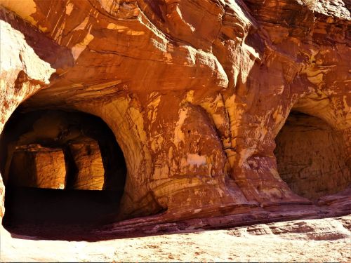 Two large, natural rock formations with smooth, reddish-brown surfaces and dark openings, set in a sandy environment.