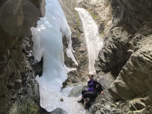 A person sits on rocks near a waterfall in a narrow canyon, surrounded by ice and steep stone walls.