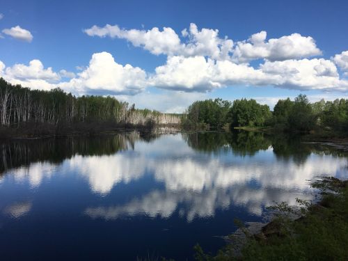 A serene lake reflecting fluffy clouds and lush greenery under a bright blue sky.