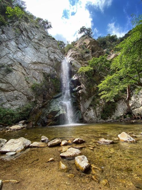 A serene waterfall cascading down rocky cliffs into a clear pool, surrounded by lush greenery and a blue sky.