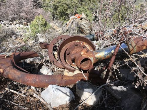 Rusty metal pipes and machinery parts are tangled in dry brush and rocks, surrounded by sparse vegetation.