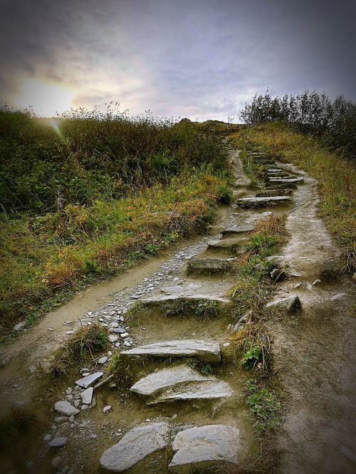 A winding stone path leads up a grassy hill under a cloudy sky, surrounded by greenery.