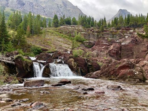 A serene waterfall cascades over rocky terrain, surrounded by lush green trees and mountains under a cloudy sky.