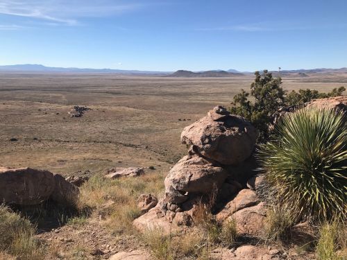 A rocky landscape with sparse vegetation, rolling hills, and a clear blue sky in the background.