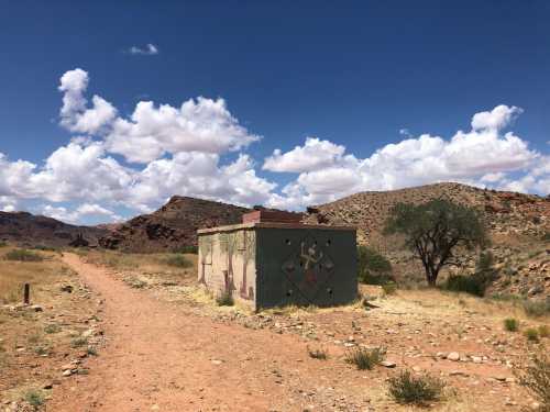 A small, weathered building on a dirt path, surrounded by rocky hills and a blue sky with fluffy clouds.