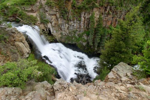 A cascading waterfall flows over rocky cliffs, surrounded by lush green trees and vegetation.