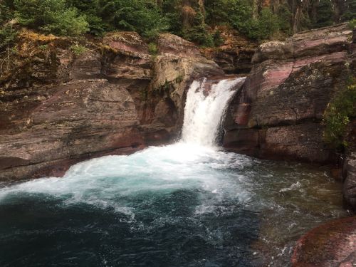 A serene waterfall cascades over rocky cliffs into a clear, turquoise pool surrounded by lush greenery.