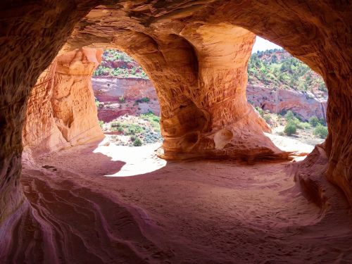 A view from inside a sandstone cave, showcasing colorful rock formations and sunlight illuminating the sandy floor.