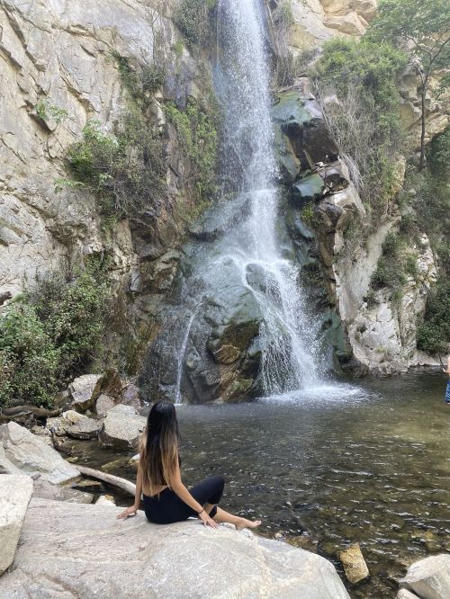 A person sits by a waterfall, surrounded by rocky terrain and lush greenery, enjoying the serene natural setting.