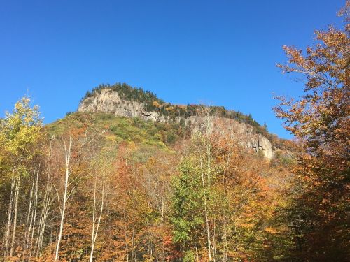 A rocky mountain peak surrounded by vibrant autumn foliage under a clear blue sky.
