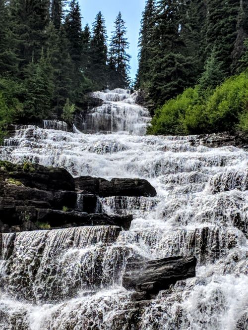 A cascading waterfall flows down rocky steps, surrounded by lush green trees and a clear blue sky.