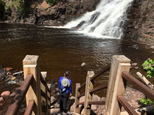 A person stands on wooden steps overlooking a waterfall and a serene pool of water surrounded by rocks and greenery.