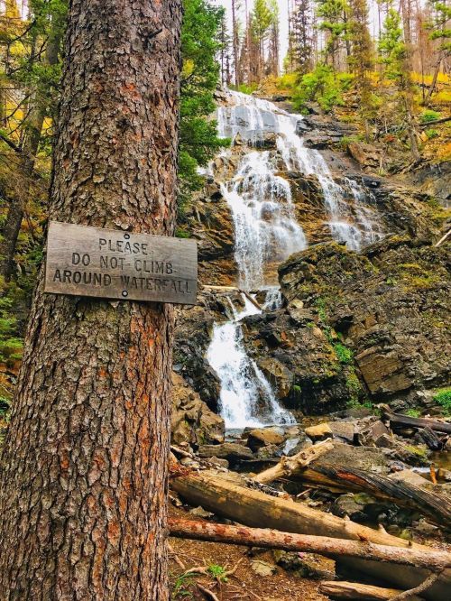 A waterfall cascades down rocky terrain, with a warning sign on a tree urging visitors not to climb around it.
