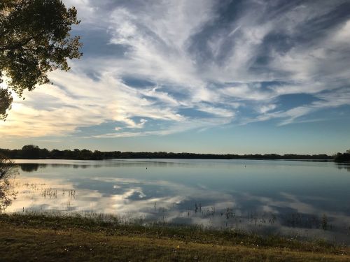 A serene lake reflecting a blue sky with wispy clouds, surrounded by trees and grassy shoreline.