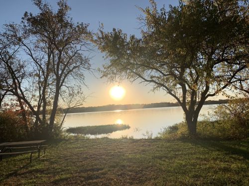 Sunrise over a calm lake, framed by trees and a grassy area, with a picnic table in the foreground.