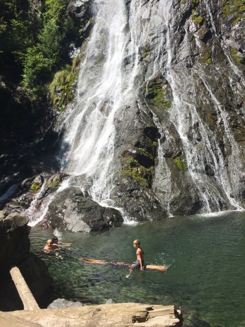 Two people swim in a natural pool near a waterfall, surrounded by lush greenery and rocky terrain.
