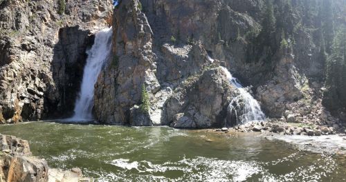 A scenic view of two waterfalls cascading down rocky cliffs into a green river surrounded by trees.