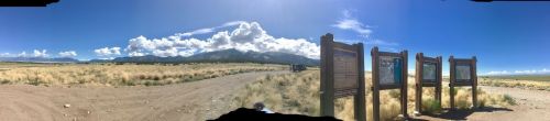 Panoramic view of a grassy landscape with mountains in the background and informational signs along a dirt road.