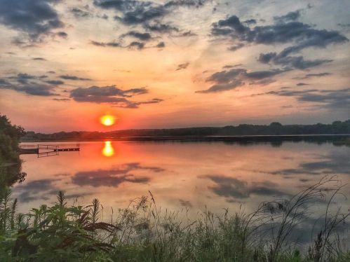 A serene sunset over a calm lake, with vibrant orange and pink hues reflecting on the water and clouds in the sky.