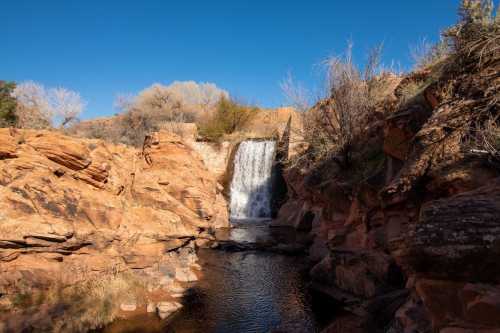 A serene waterfall cascades over rocky cliffs into a calm pool, surrounded by dry vegetation and clear blue skies.