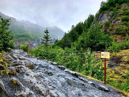 A rocky trail leads through lush greenery, with mountains shrouded in mist and a warning sign nearby.