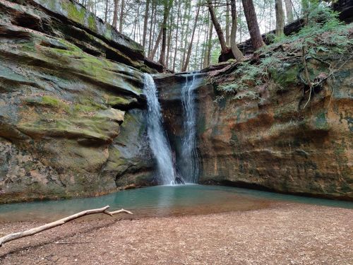 A serene waterfall cascades into a clear pool, surrounded by mossy rocks and lush green trees.